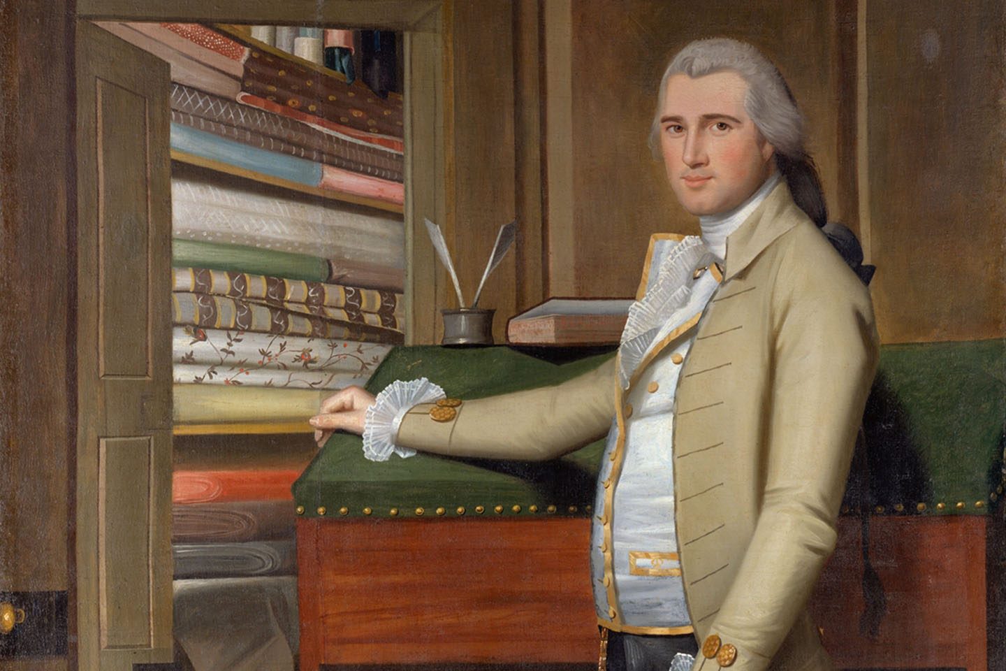 A man dressed in colonial attire rests his hand on a writing desk adjacent to an open door that looks into a room of shelves storing an abundance of folded textiles